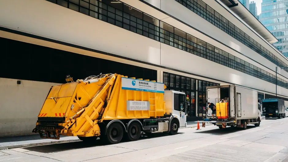 A vibrant urban street scene showing garbage and delivery trucks beside a modern building.
