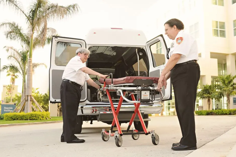 Two emergency responders load a stretcher into an ambulance outdoors, depicting medical assistance.